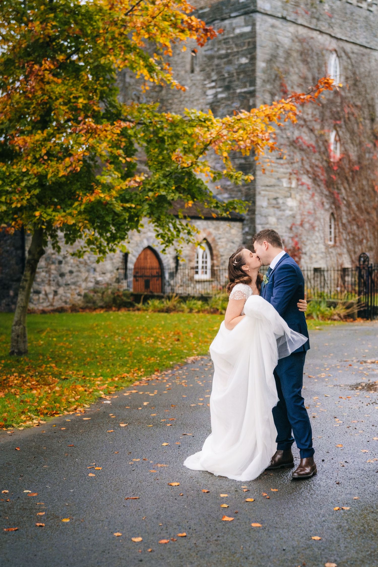 Newlyweds share romantic moments against autumn foliage and stone church backdrop.