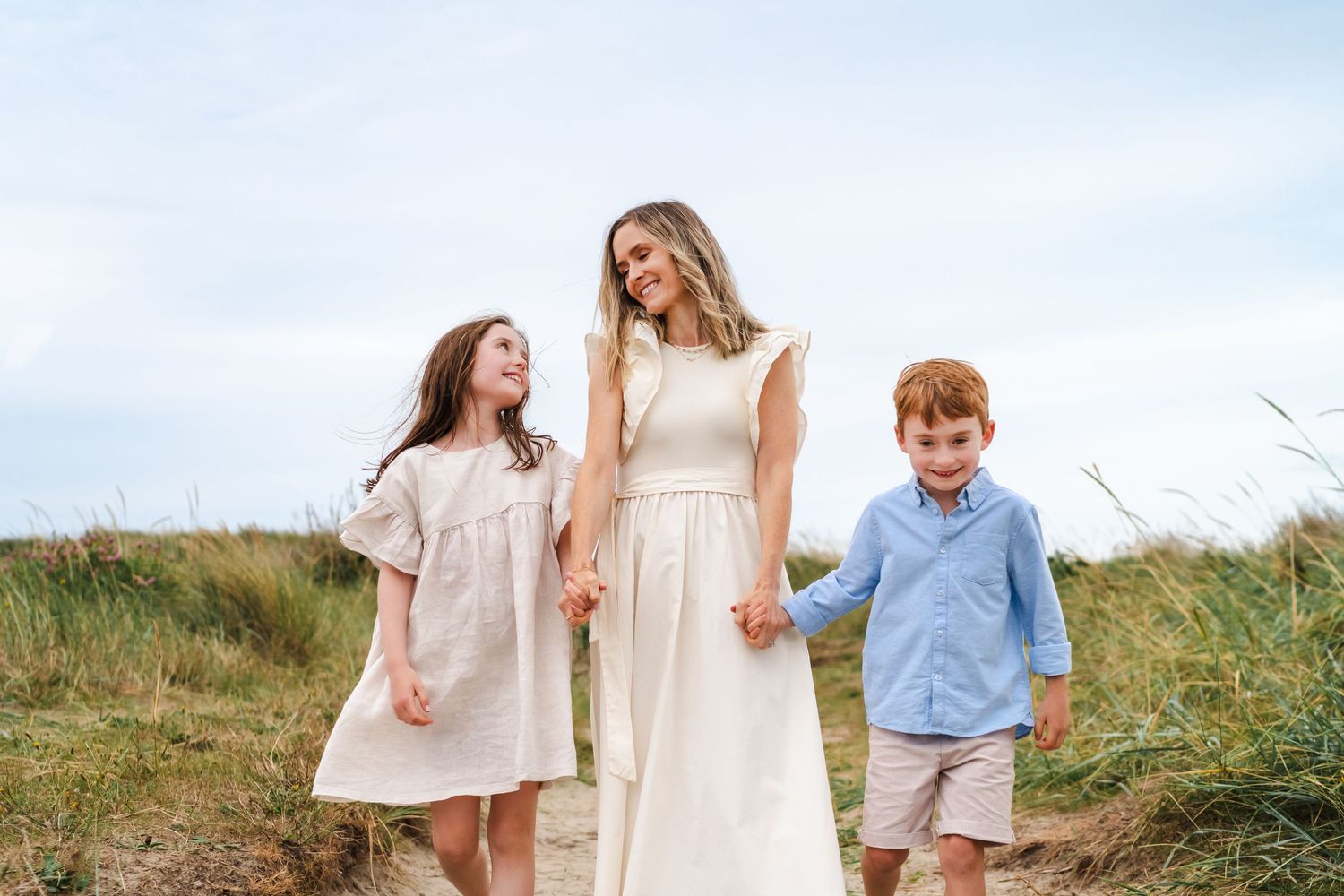 Family walking together through tall beach grass on a sunny day, with white and blue summer outfits.  Family photographer Dublin Ireland