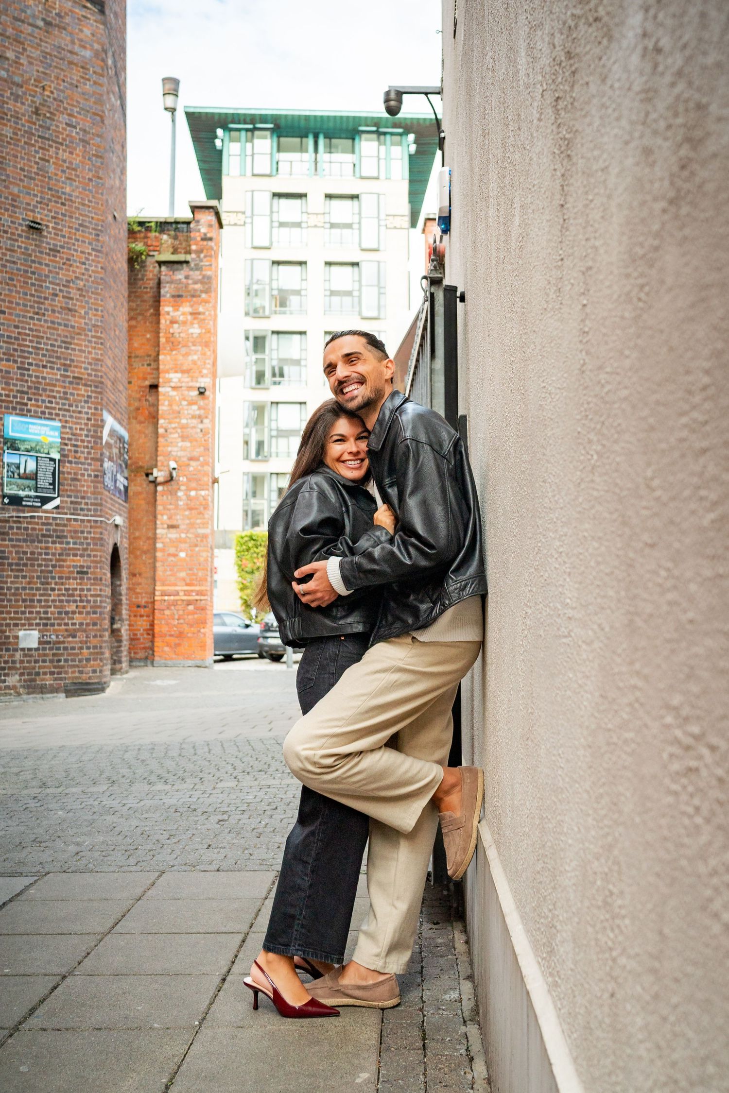 A couple shares a romantic moment against a brick wall in an urban setting, dressed in casual chic attire. Couple's Photoshoot PHOTOGRAPHER