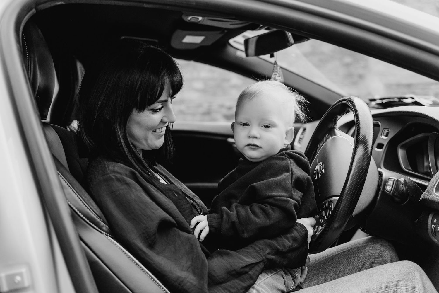 A parent and baby sit in the driver's seat of a car together in black and white.  Family PHOTOGRAPHER Dublin