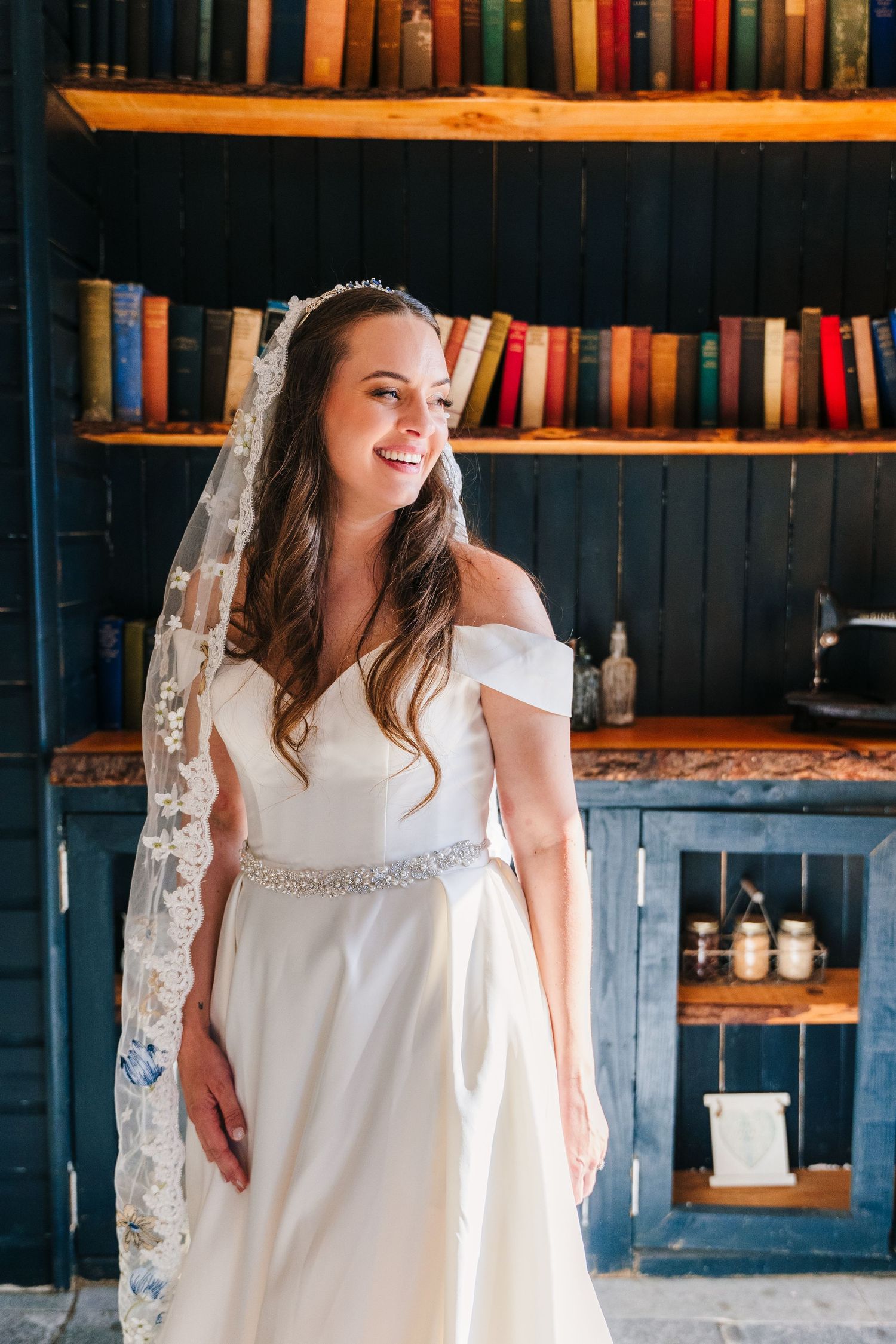 Poulaphouca Wedding photograpBeautiful wedding gown with off-shoulder design and lace veil photographed against rustic bookshelf background.