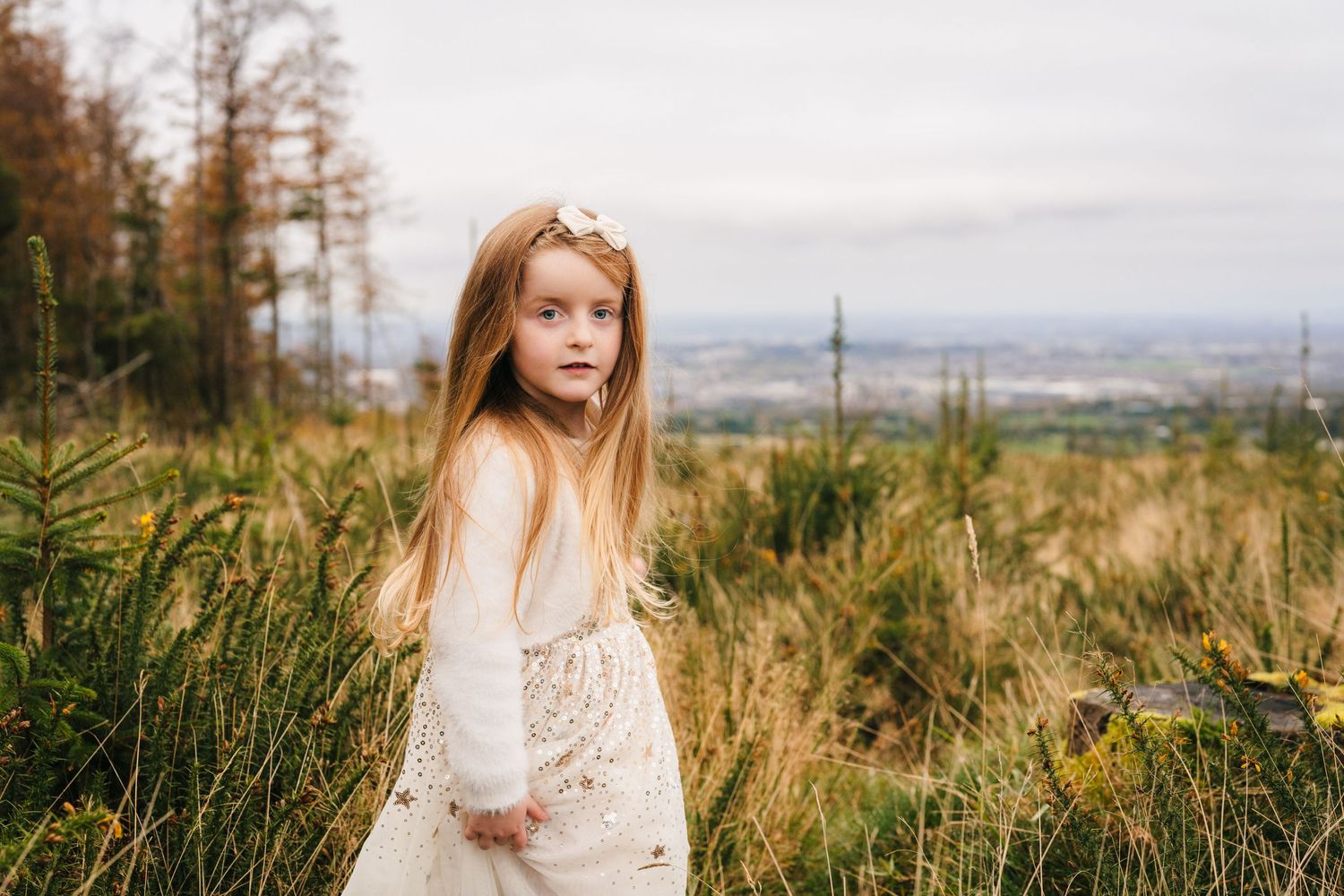 Family Photographer Dublin Ireland A young person in a white dress stands among tall grass with a scenic valley view in the background.
