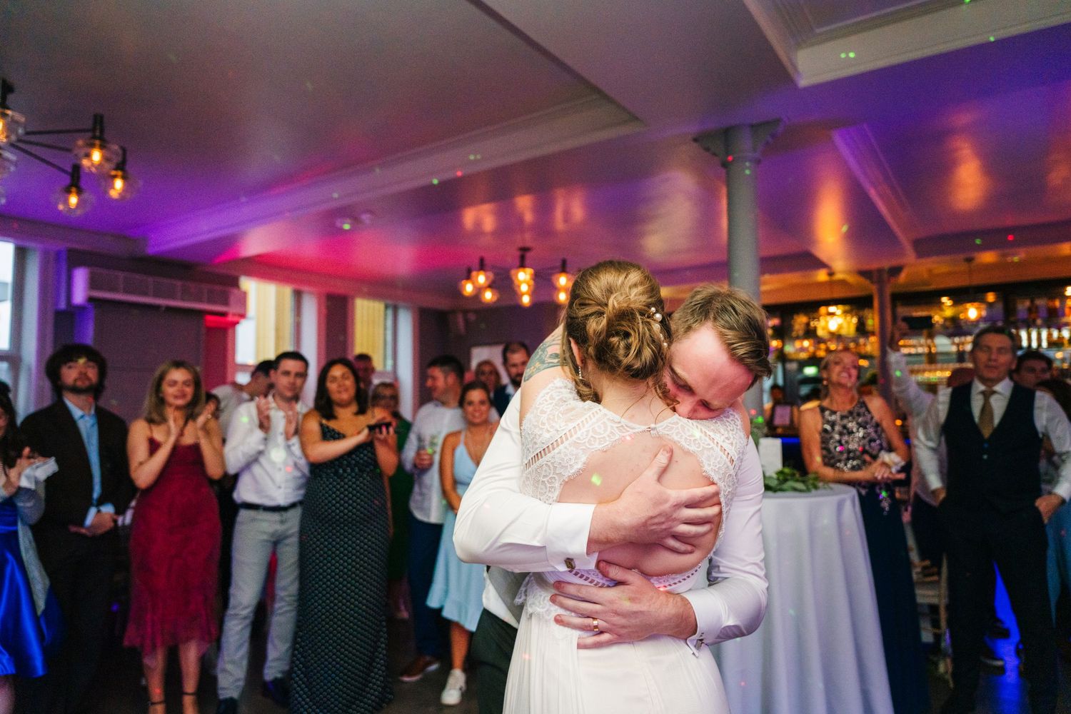 Wedding couple share their first dance in a venue with purple uplighting while guests watch at cocktail tables. PHOTOGRAPHER Dublin Ireland