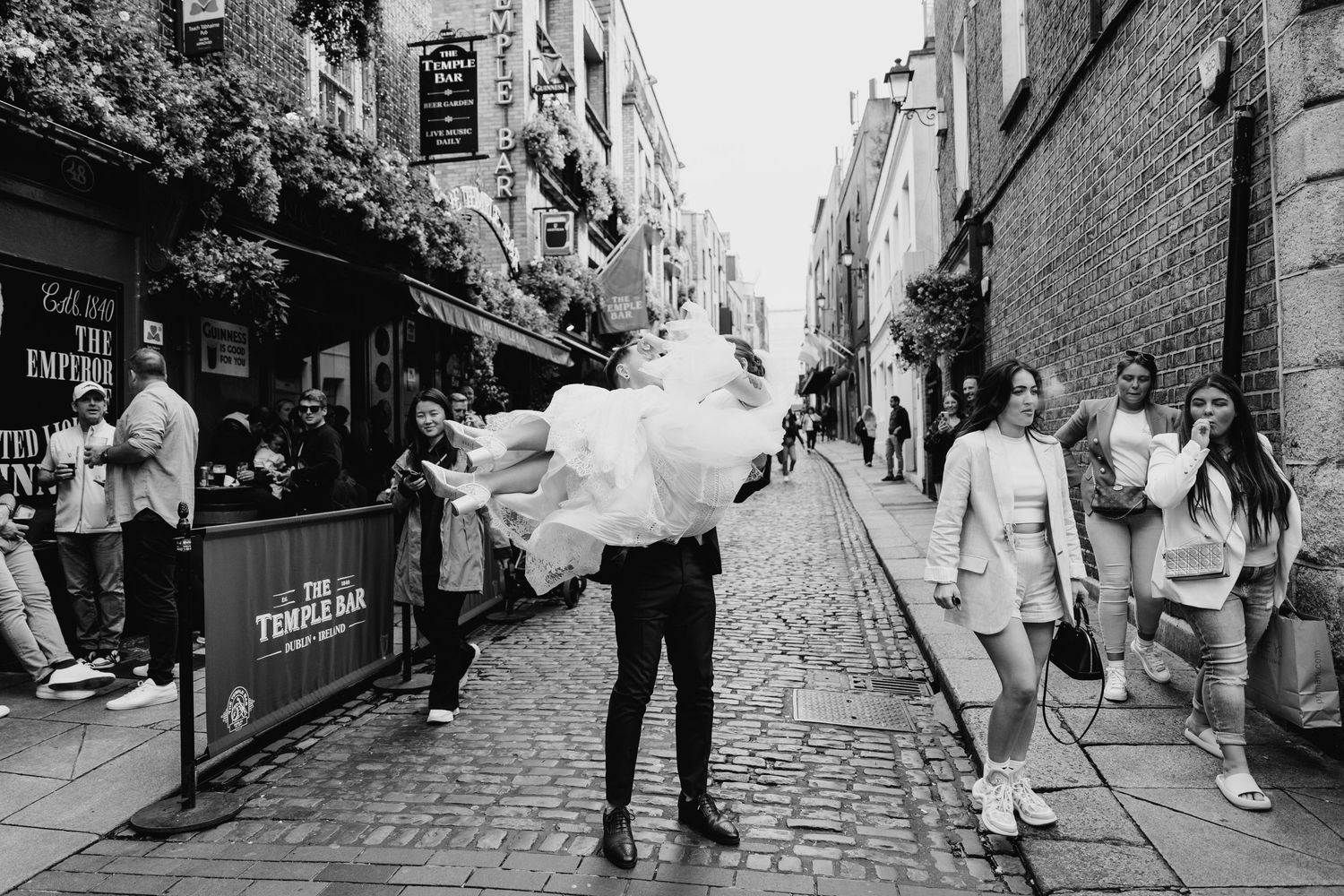 A person carries a bride in a flowing white dress through a cobblestone street while pedestrians look on. PHOTOGRAPHER Dublin Ireland Weddin