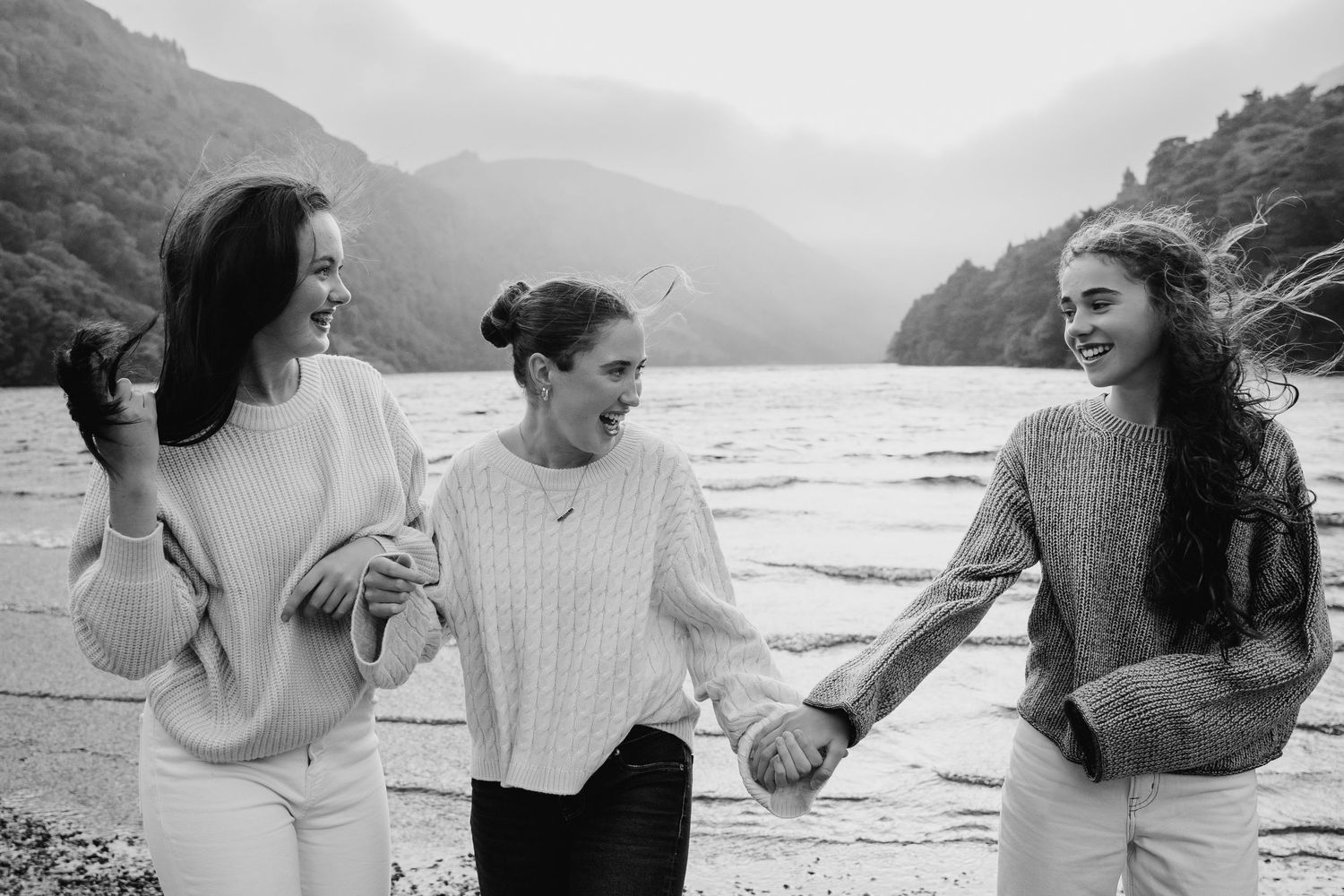 Family photographer Ireland Dublin Friends in sweaters sharing candid laughs against a scenic mountain lake backdrop.