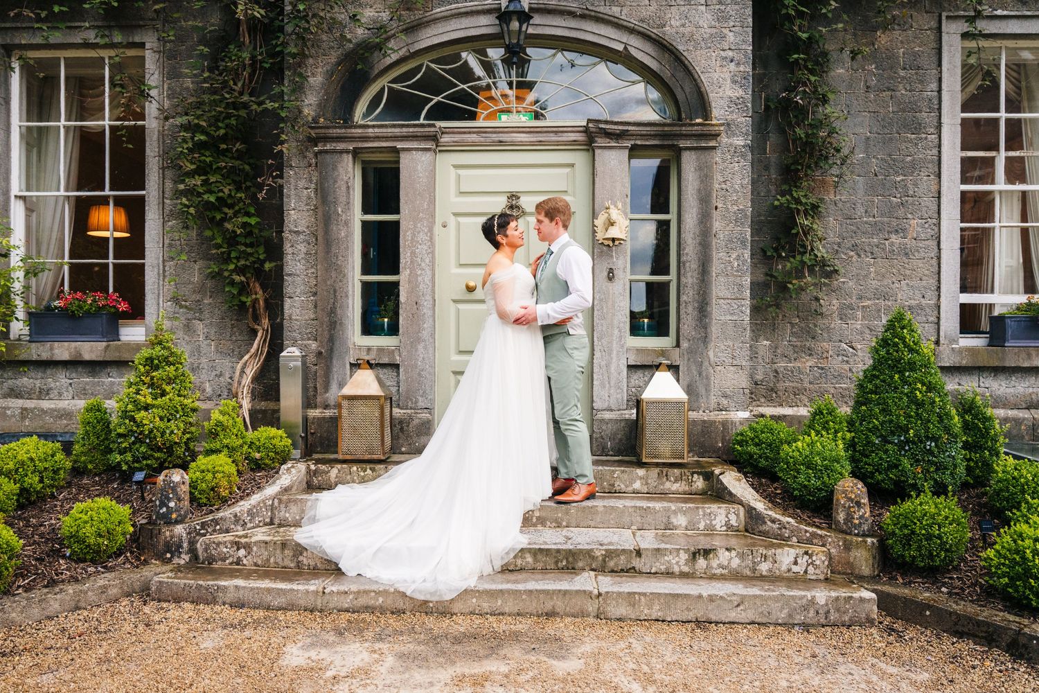 Millhouse slane wedding phWedding couple shares intimate moment on stone steps of elegant mansion with arched doorway and manicured gardens.