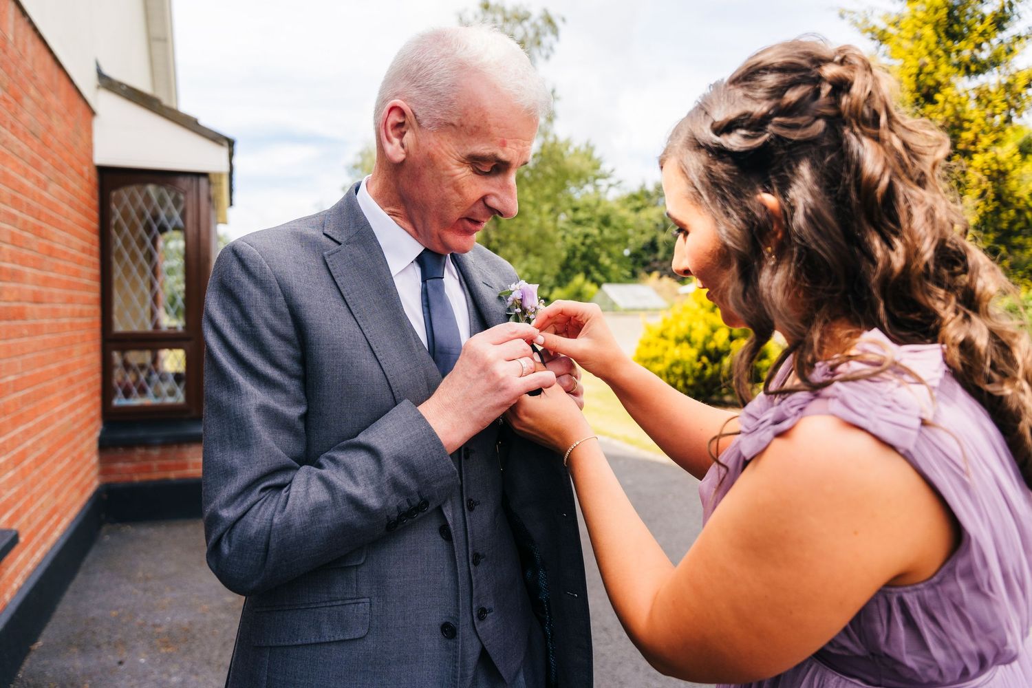 Two people share an intimate moment while pinning a boutonniere onto a formal grey suit at an outdoor event. Millhouse slane wedding photogr