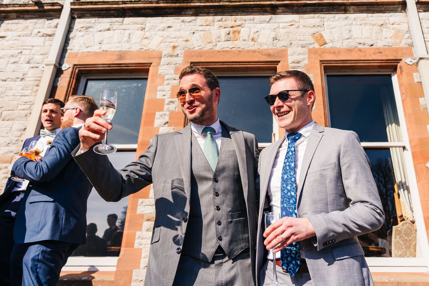 Castle Leslie Wedding Photographer Dublin Group of wedding guests in suits enjoying drinks and conversation outside brick building.