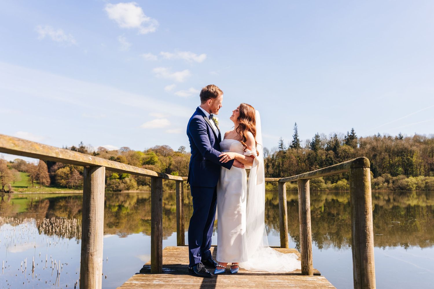 Newlywed couple stands together on a wooden dock overlooking a serene lake on their wedding day. Castle Leslie Wedding Photographer Dublin