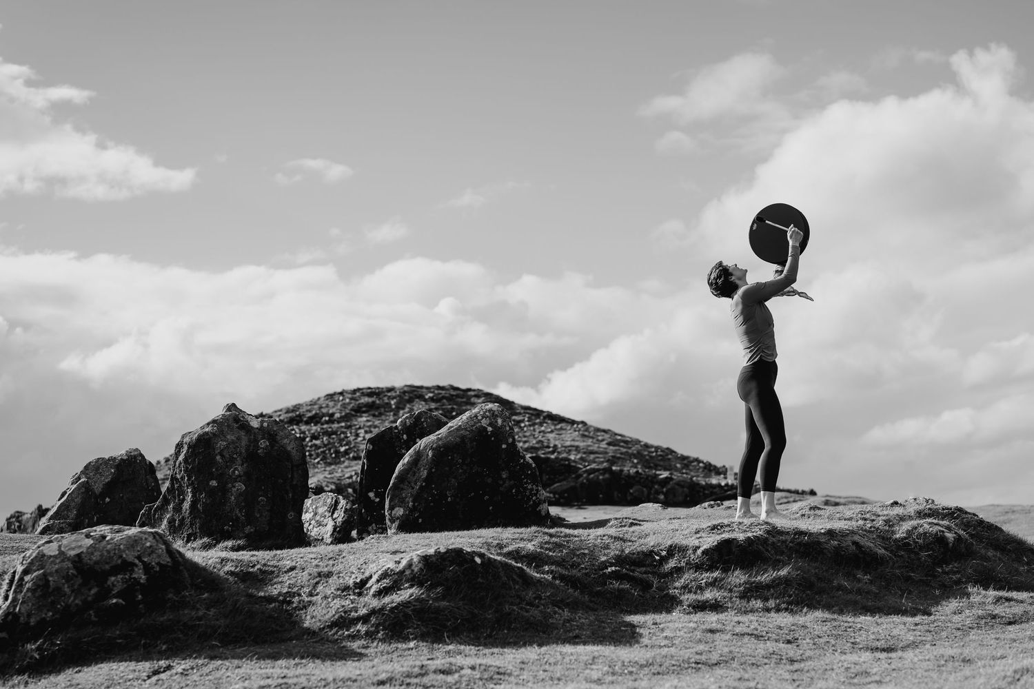 A silhouette stands atop a rocky outcrop holding a round object overhead against a cloudy black and white sky. Portrait PHOTOGRAPHER Dublin