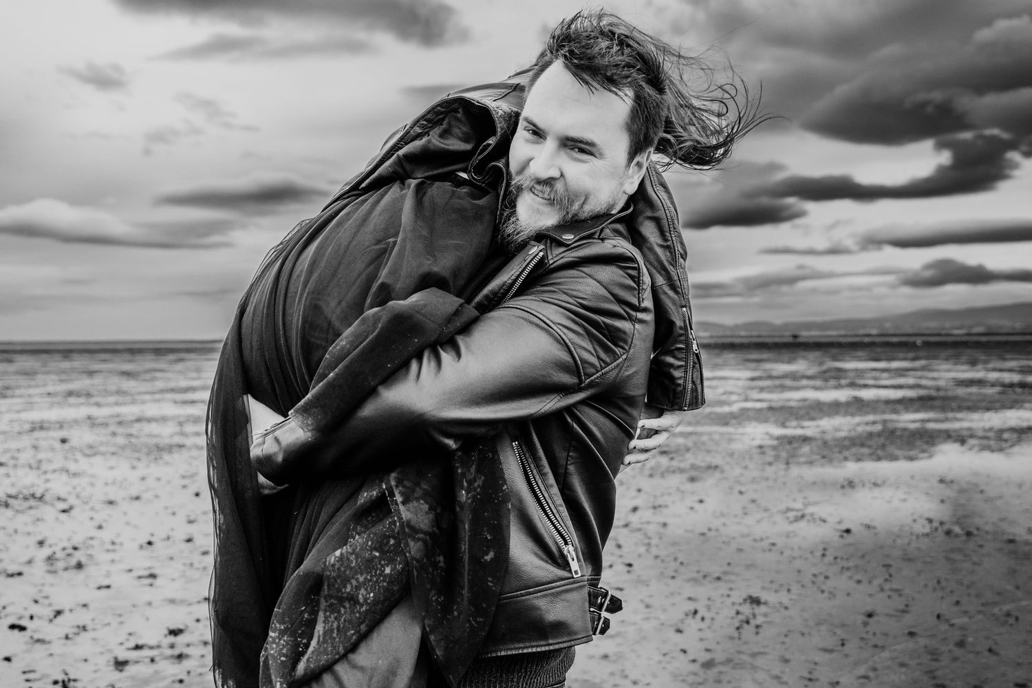 Intimate black and white portrait of couple embracing on windswept beach. engagement PHOTOGRAPHER Dublin