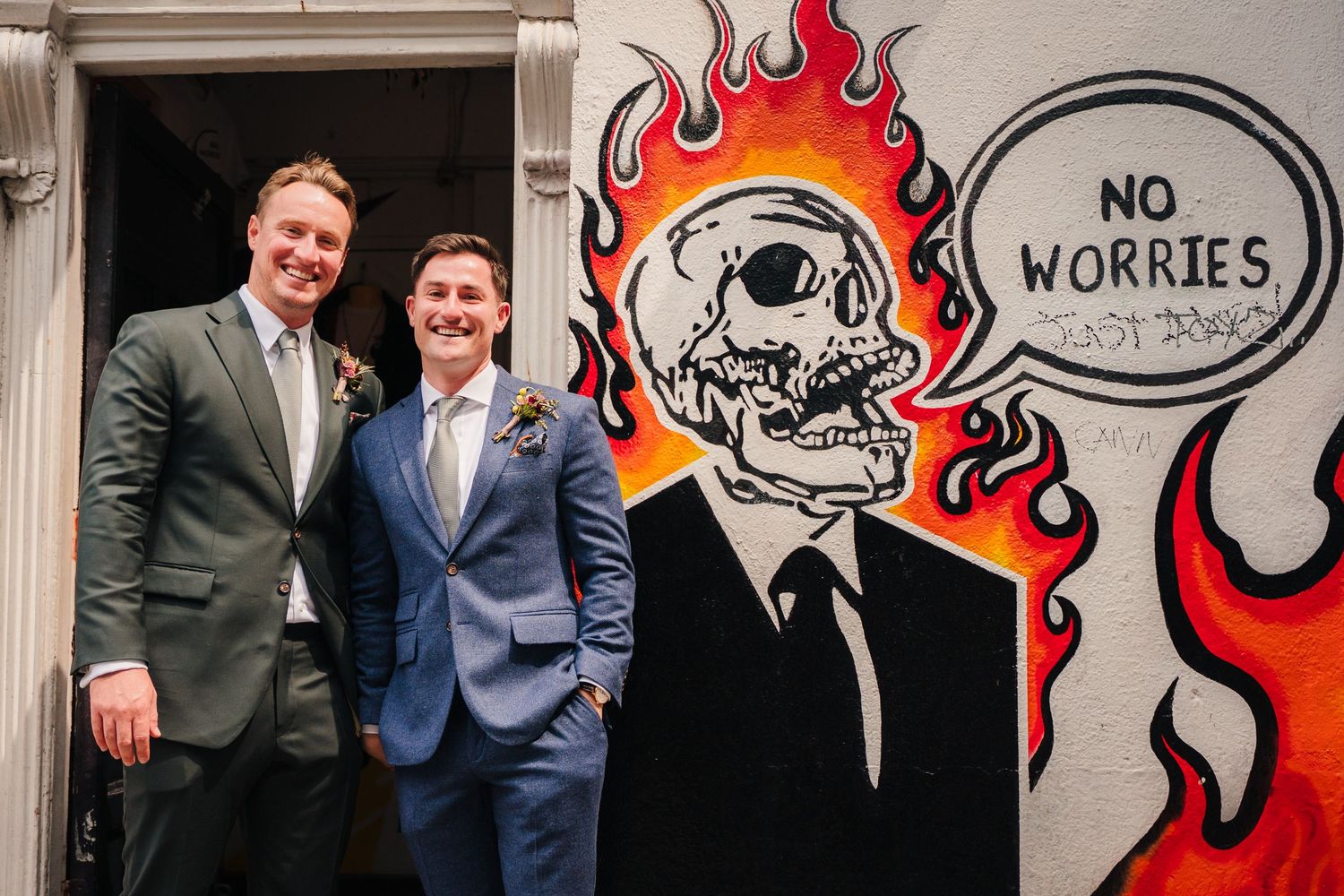 Wedding party poses in front of vibrant graffiti wall with flames and cartoon artwork at outdoor venue. Same sex wedding PHOTOGRAPHER Dublin