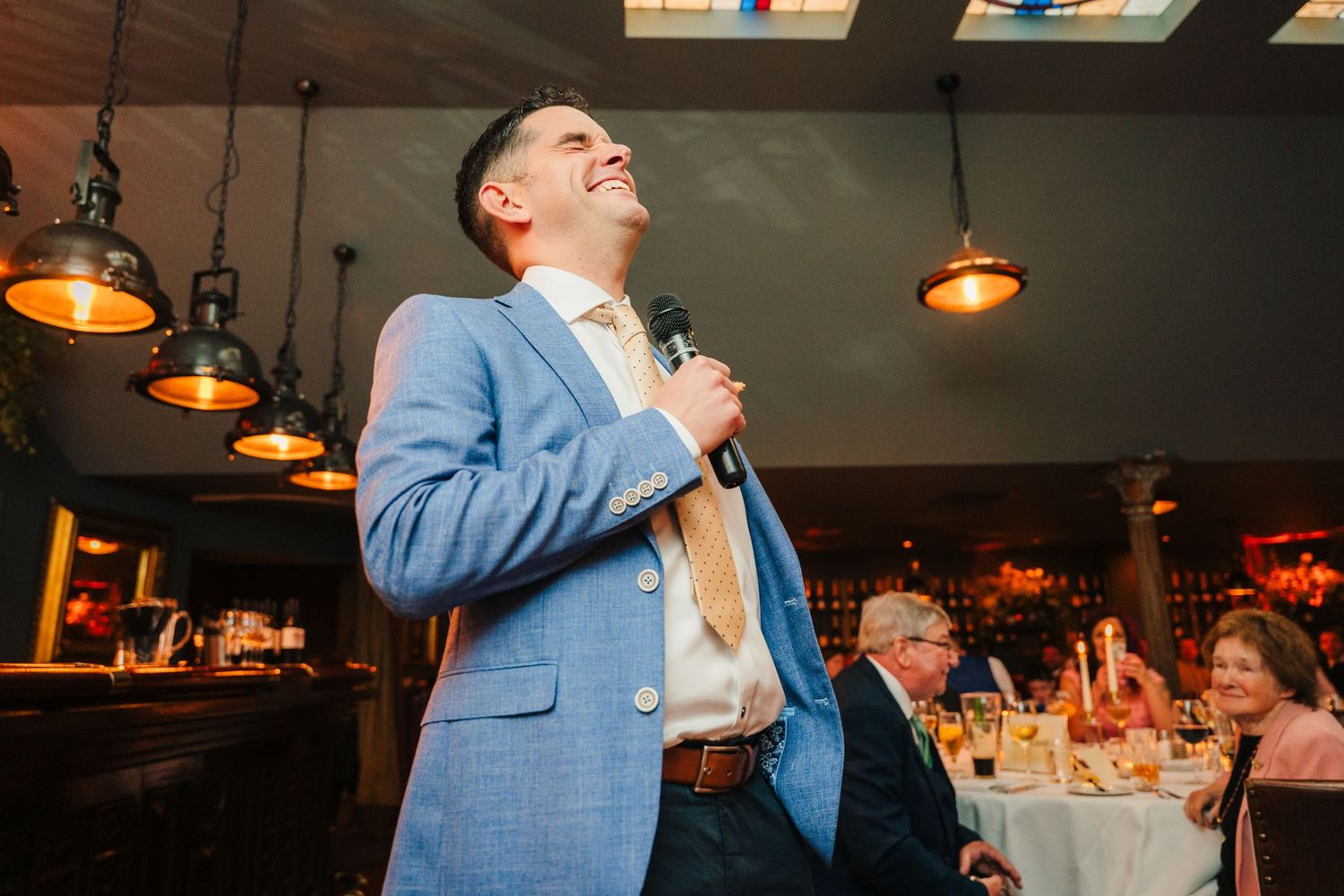 A person in a light blue suit gives a speech at a wedding reception under pendant lighting. Marco Pierre Dublin Wedding Photographer