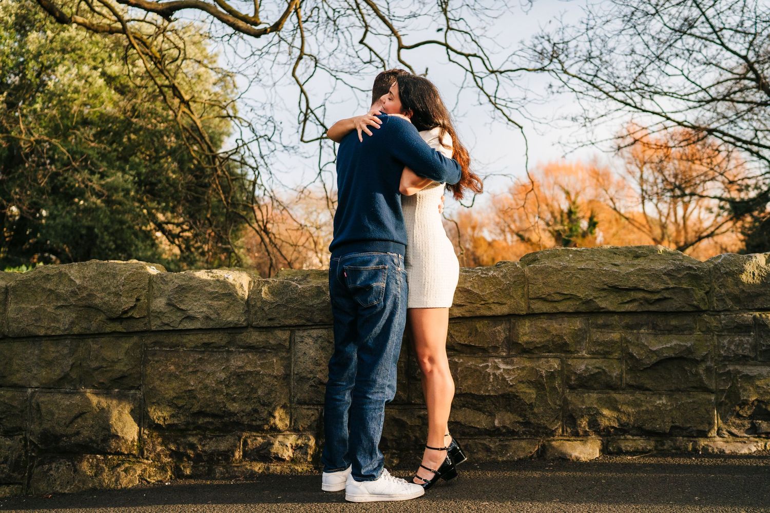 Silhouettes stand close together against a stone wall backdrop during a golden autumn sunset. Surprise proposal dublin photography