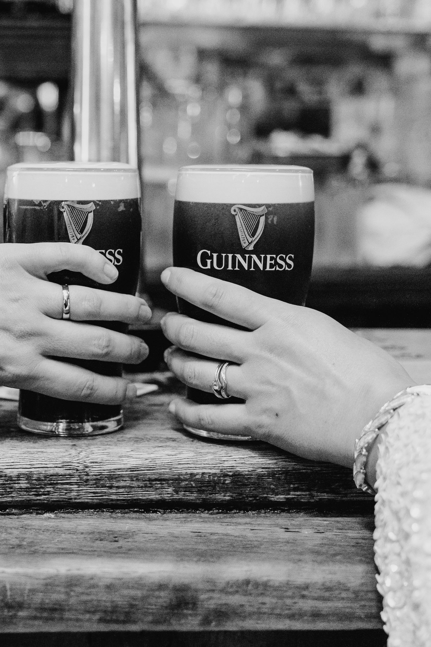 Two hands clink Guinness pint glasses together on a wooden bar counter in black and white. House Dublin Wedding Photographer