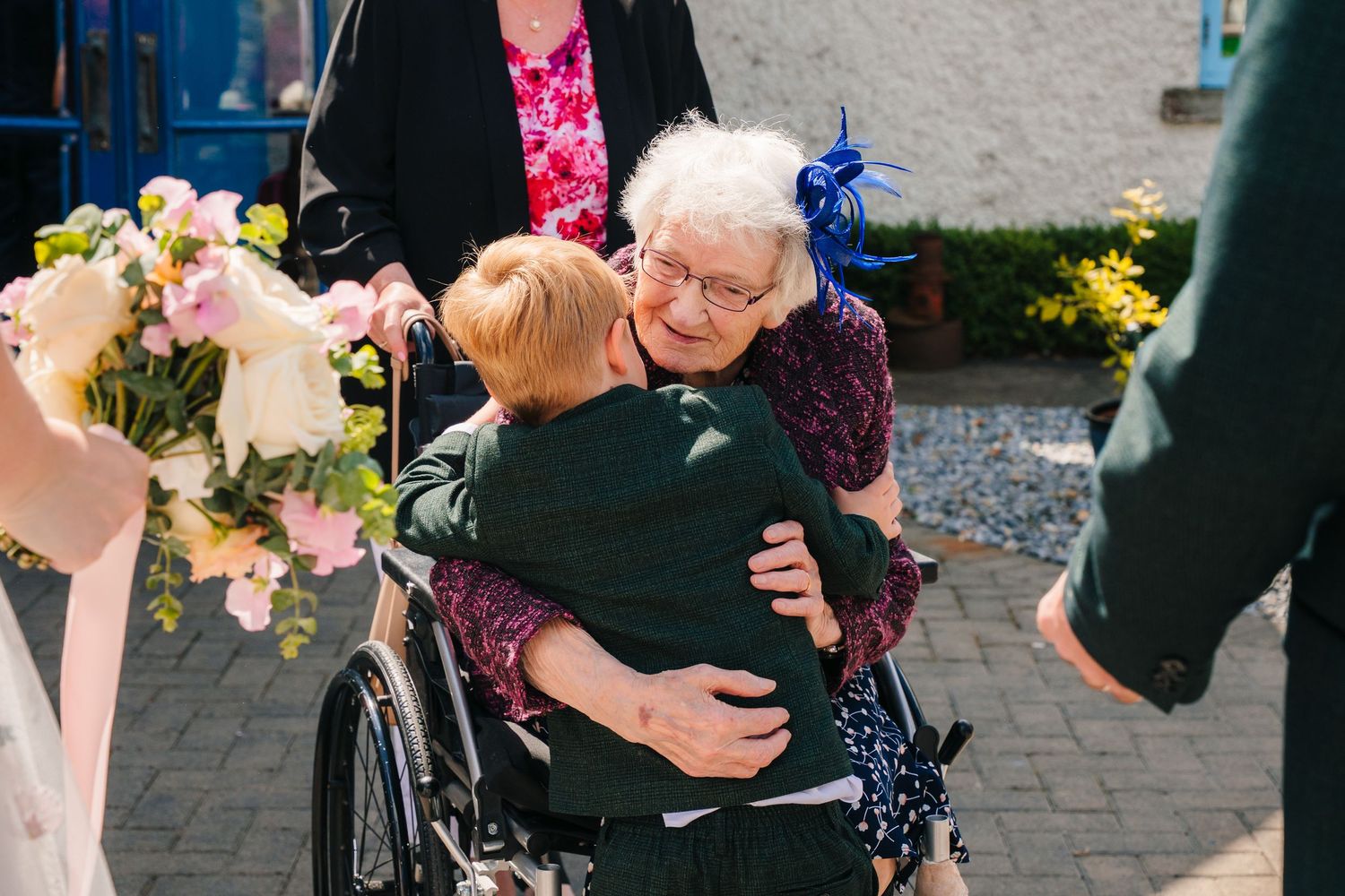 A group of wedding guests gather outdoors sharing joyful hugs and embraces on a paved patio area. . Village Barn Tyrrellspass wedding photog