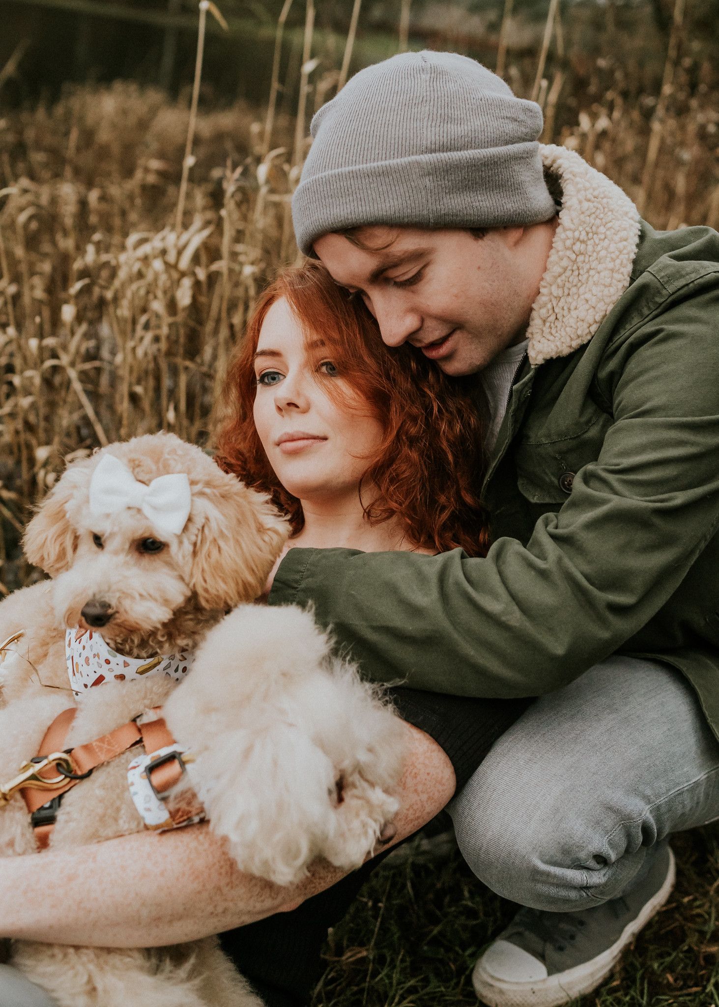 A couple cuddles with their fluffy white dog while sitting in tall autumn grass during an outdoor photoshoot. PHOTOGRAPHER Dublin ireland