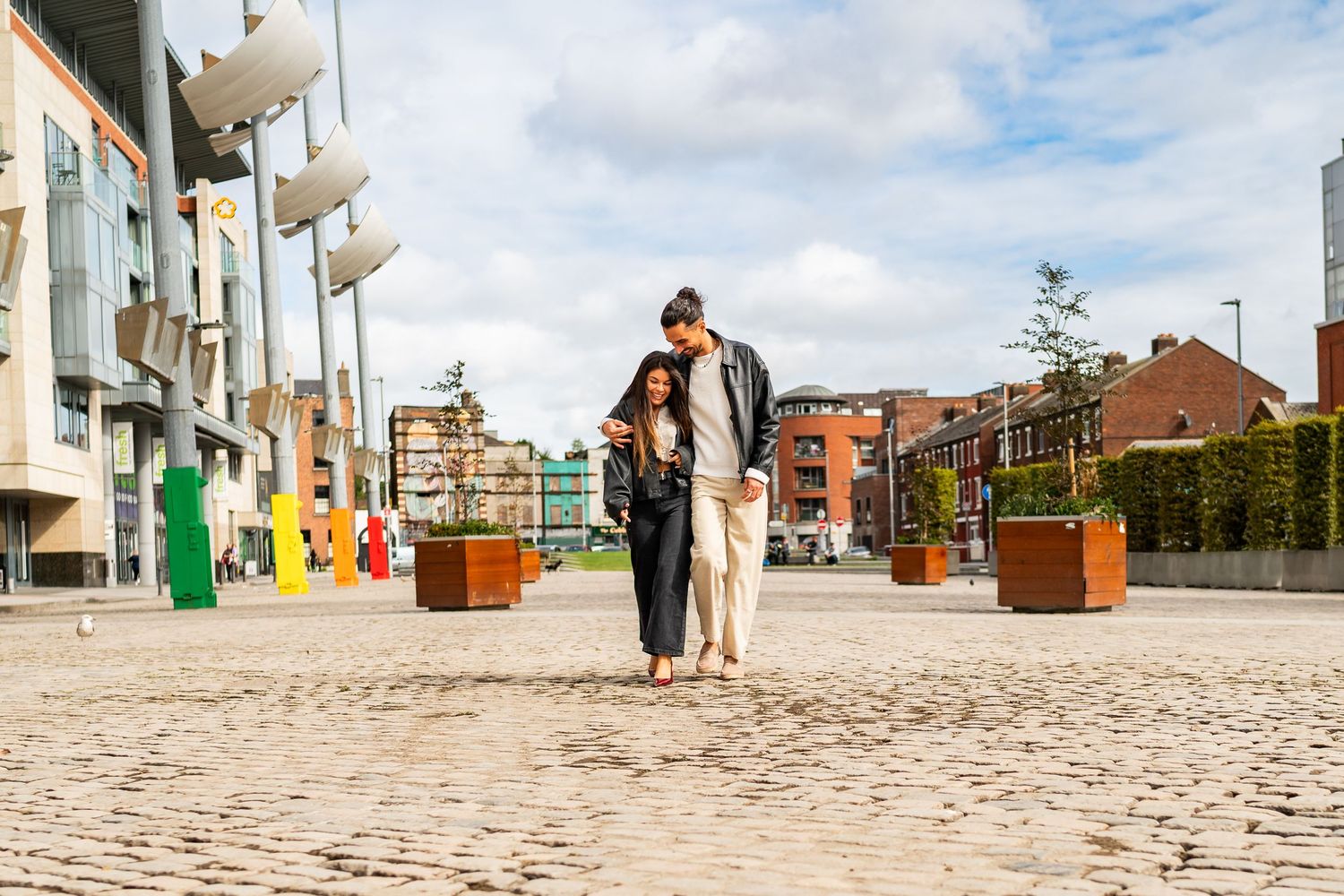People walking across a paved stone plaza with modern buildings and decorative planters in the background. Couple's Photoshoot PHOTOGRAPHER