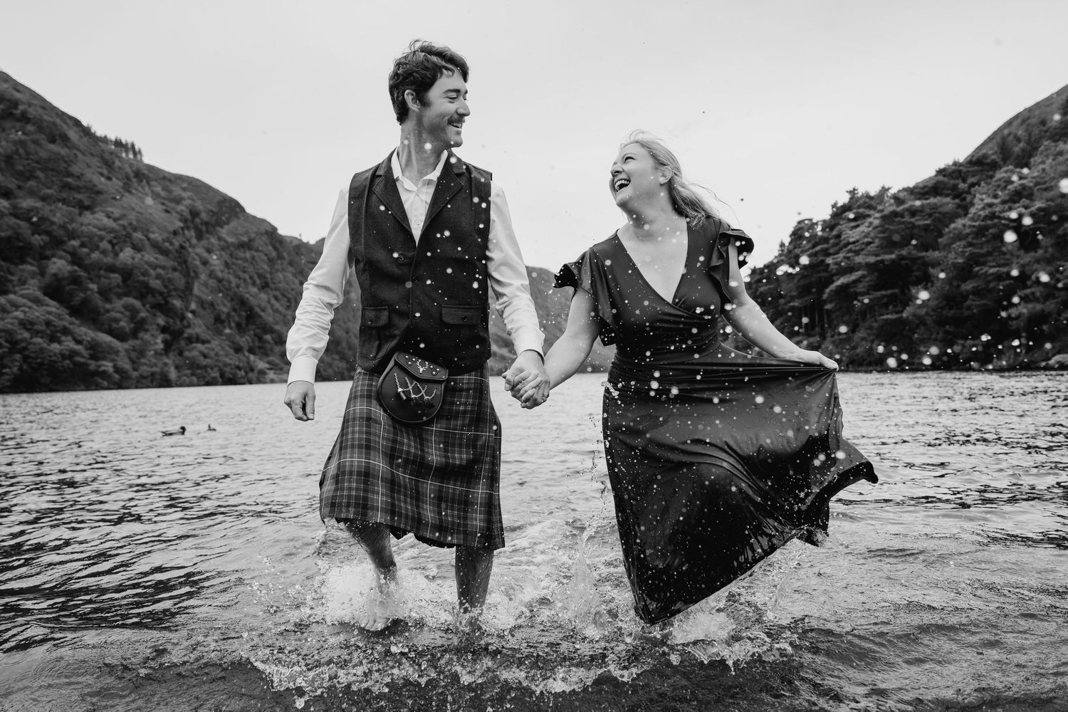 A couple splashes and plays together in the ocean water near a rocky coastline during sunset. Family photogrAPHER DUBLIN