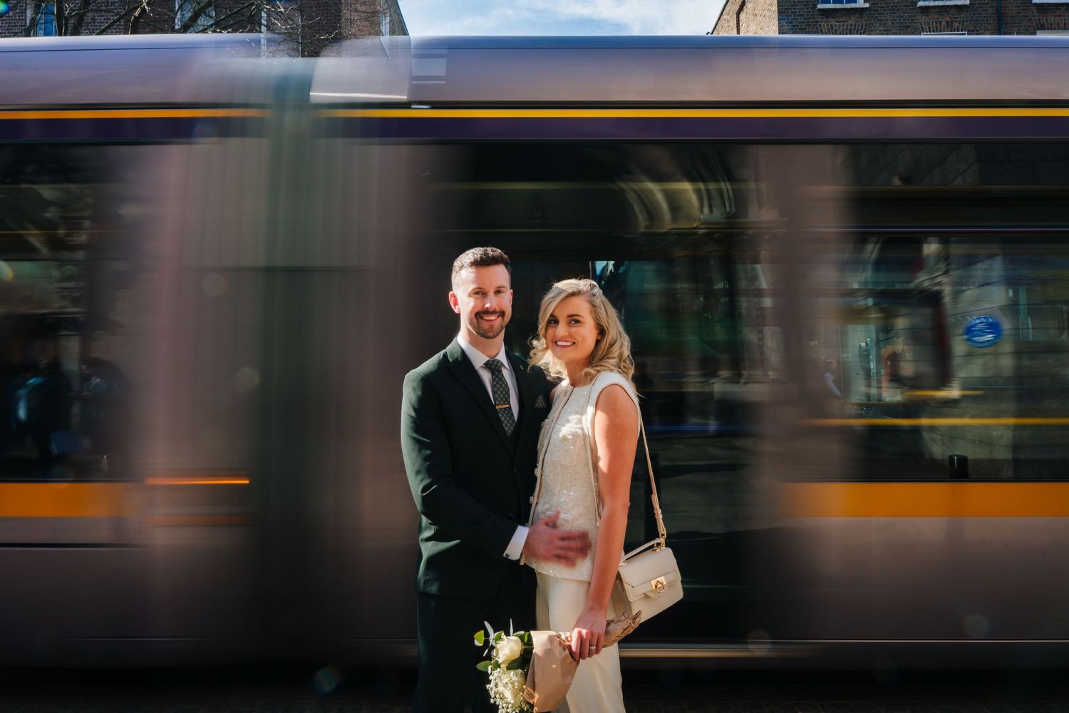 A couple stands together as a blurred train or tram passes behind them creating a dynamic motion effect. City Hall Dublin wedding photogrAPh