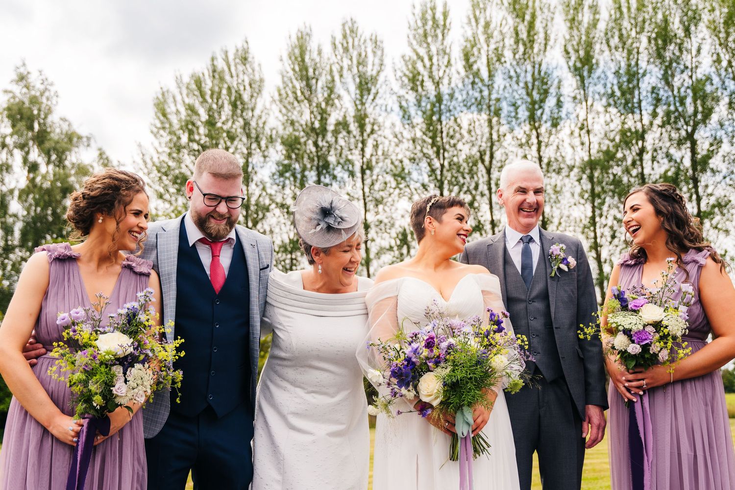 Wedding group shares joyful moment outdoors with purple bouquets and formal attire against a backdrop of tall trees.Millhouse slane wedding