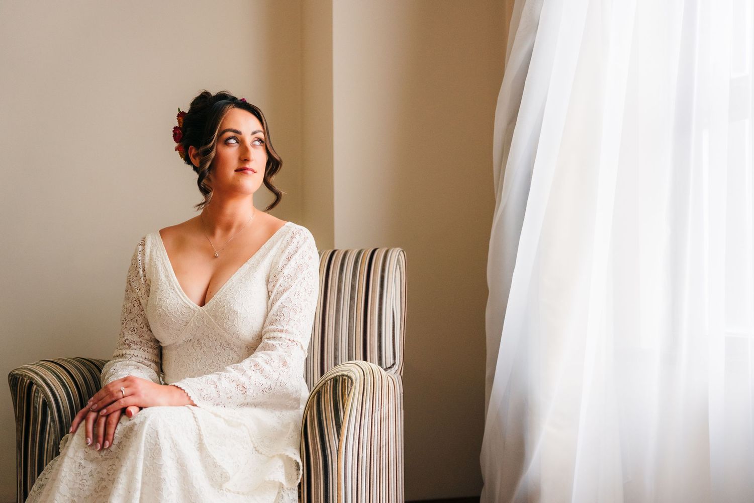 Person in elegant white lace wedding gown sits thoughtfully in striped chair by window with natural light. Wedding Photography Trim Castle I