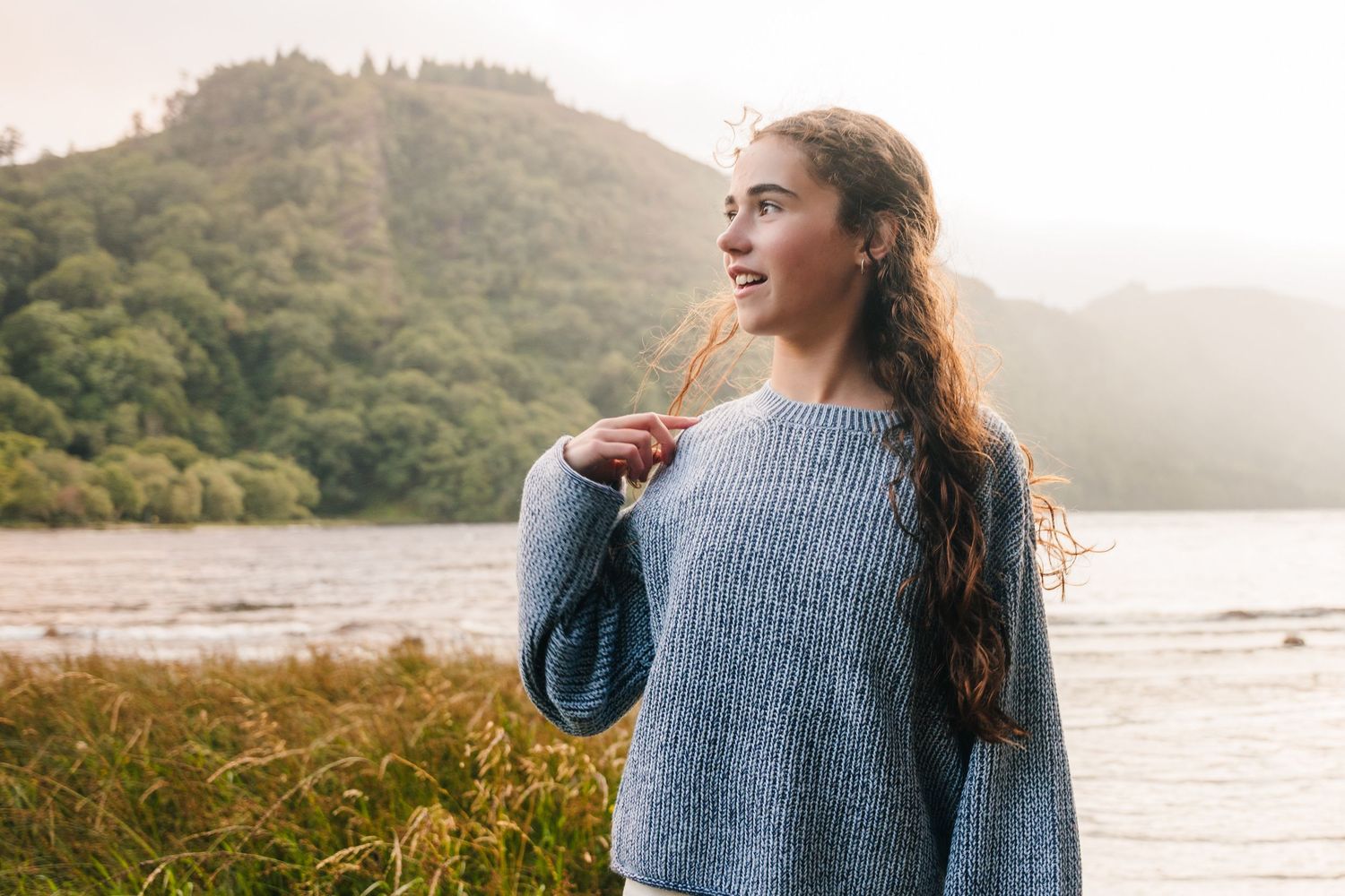 Family photographer Ireland Dublin A person in a blue sweater stands against a scenic mountain lake backdrop during golden hour.