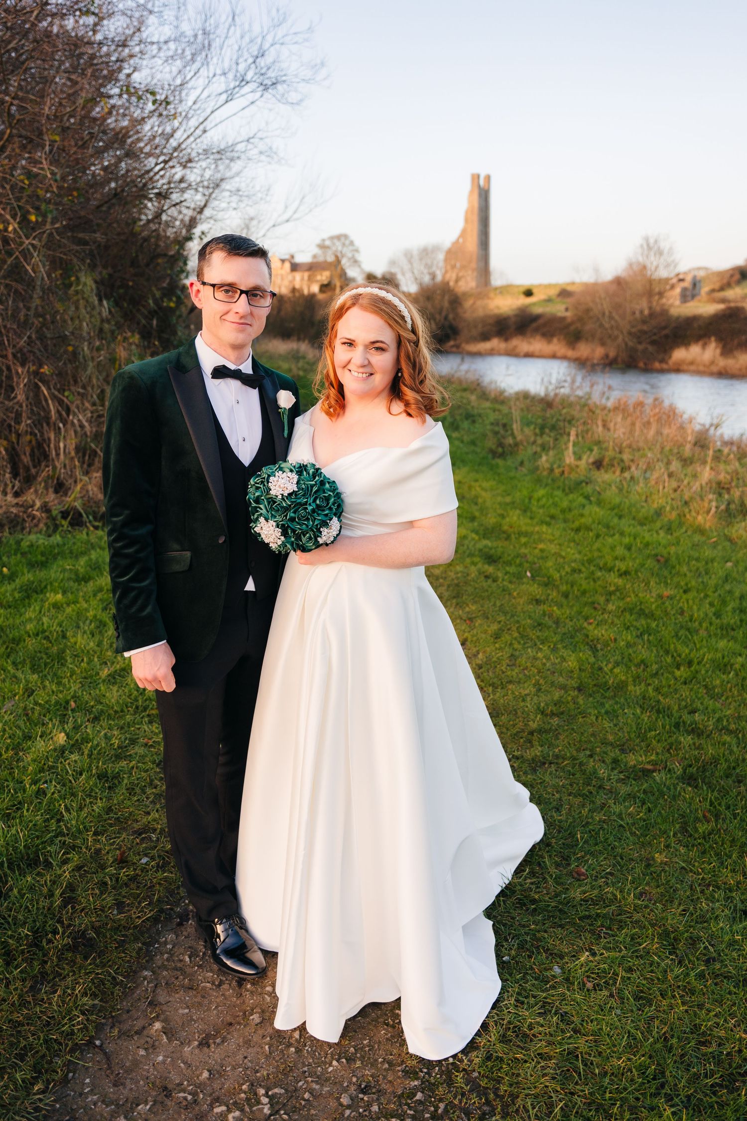 A newlywed couple poses for a portrait near a river with a church spire visible in the background.  Wedding PHOTOGRAPHER Trim Castle