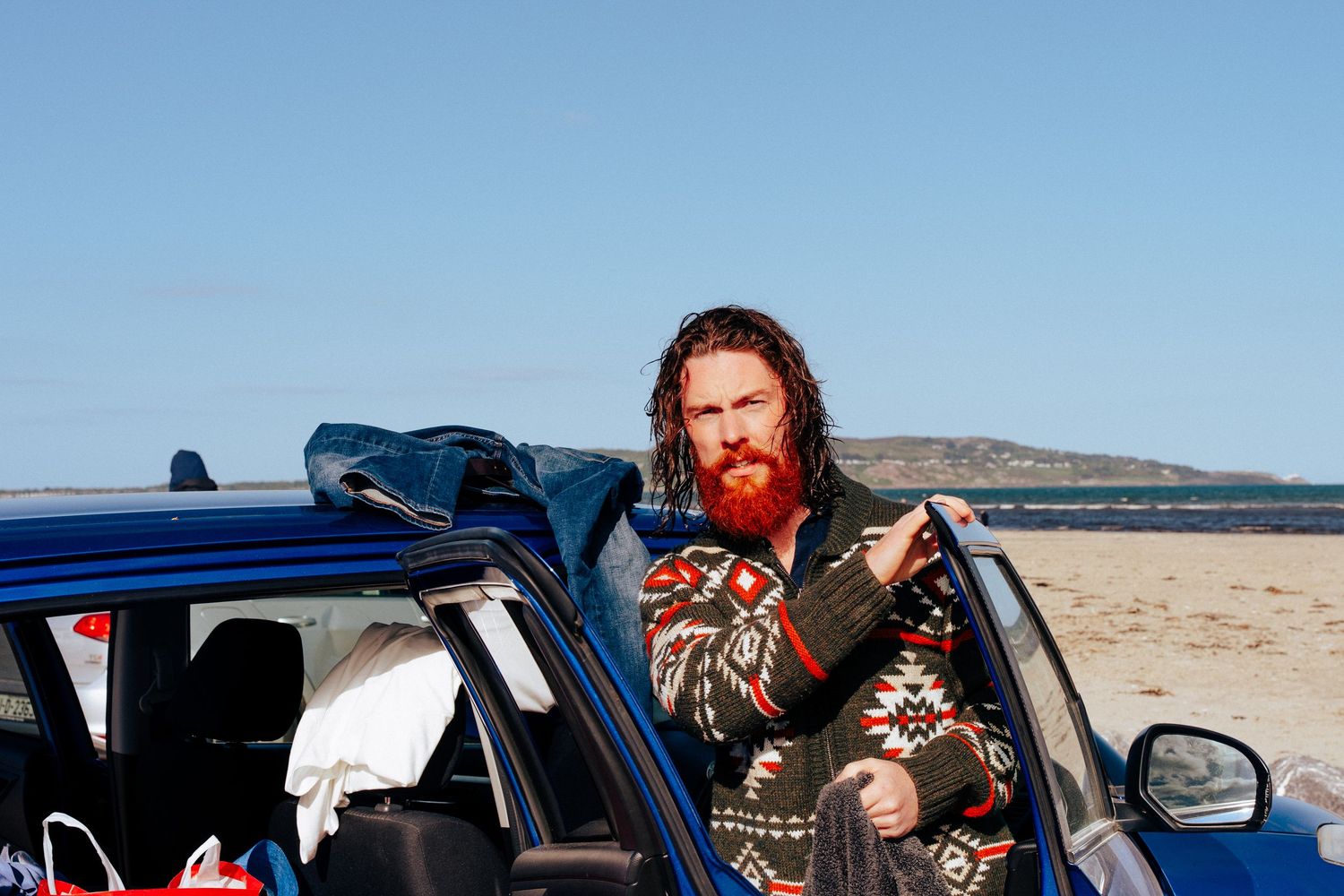 Musician with acoustic guitar posed by blue car on beach with mountains in background. PHOTOGRAPHER Dublin