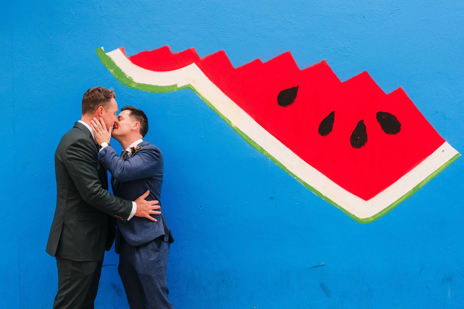Two people embrace against a vibrant blue wall with painted watermelon slice artwork. Same sex wedding PHOTOGRAPHER Dublin