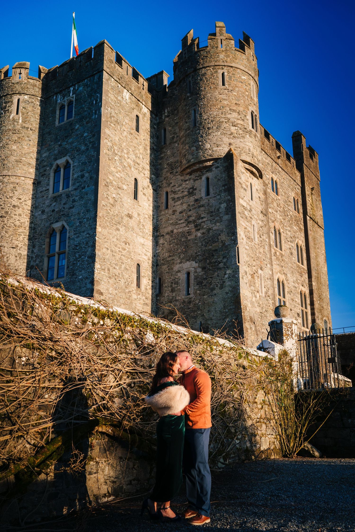 Medieval castle walls with stone turrets stand against bright blue sky as a couple shares an intimate moment. PHOTOGRAPHER Dublin ireland