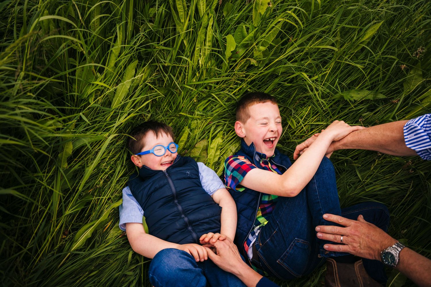 Two children lying in tall green grass reach playfully for outstretched hands on a sunny day. family PHOTOGRAPHER Dublin