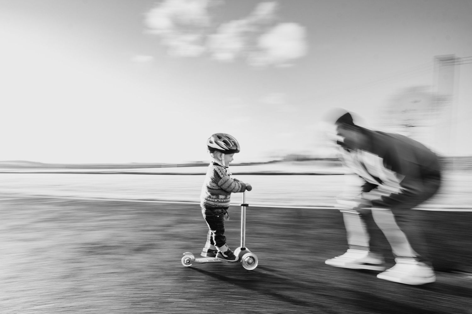 Motion-blurred black and white image of child riding scooter with adult running alongside. family PHOTOGRAPHER Dublin