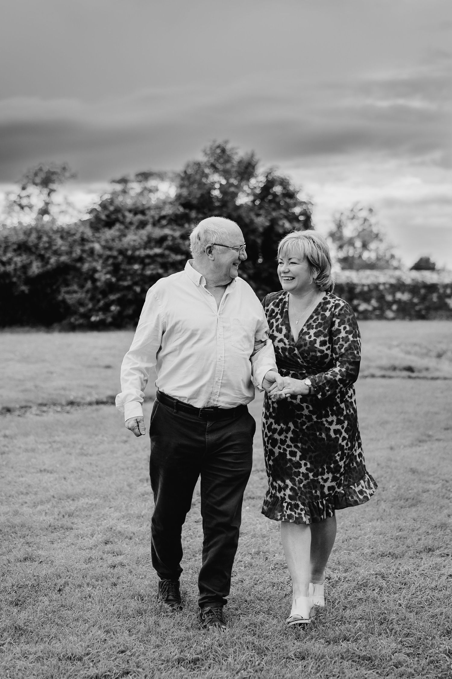 An elderly couple walks arm in arm through a grassy field on a cloudy day in black and white photography. PHOTOGRAPHER Dublin ireland