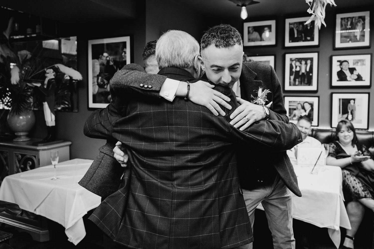 A black and white photograph captures an emotional embrace between two people at a restaurant with framed photos on the wall. Wedding PHOTOG