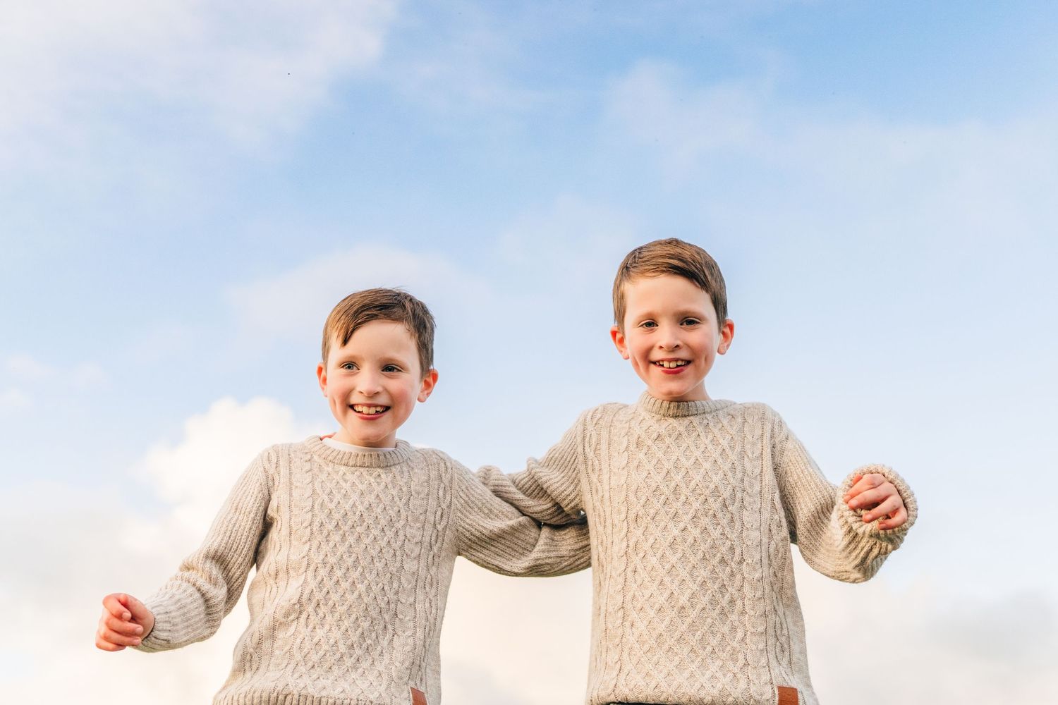 Two children wearing matching cream cable knit sweaters jump and play together against a bright cloudy sky. Family PHOTOGRAPHER Dublin