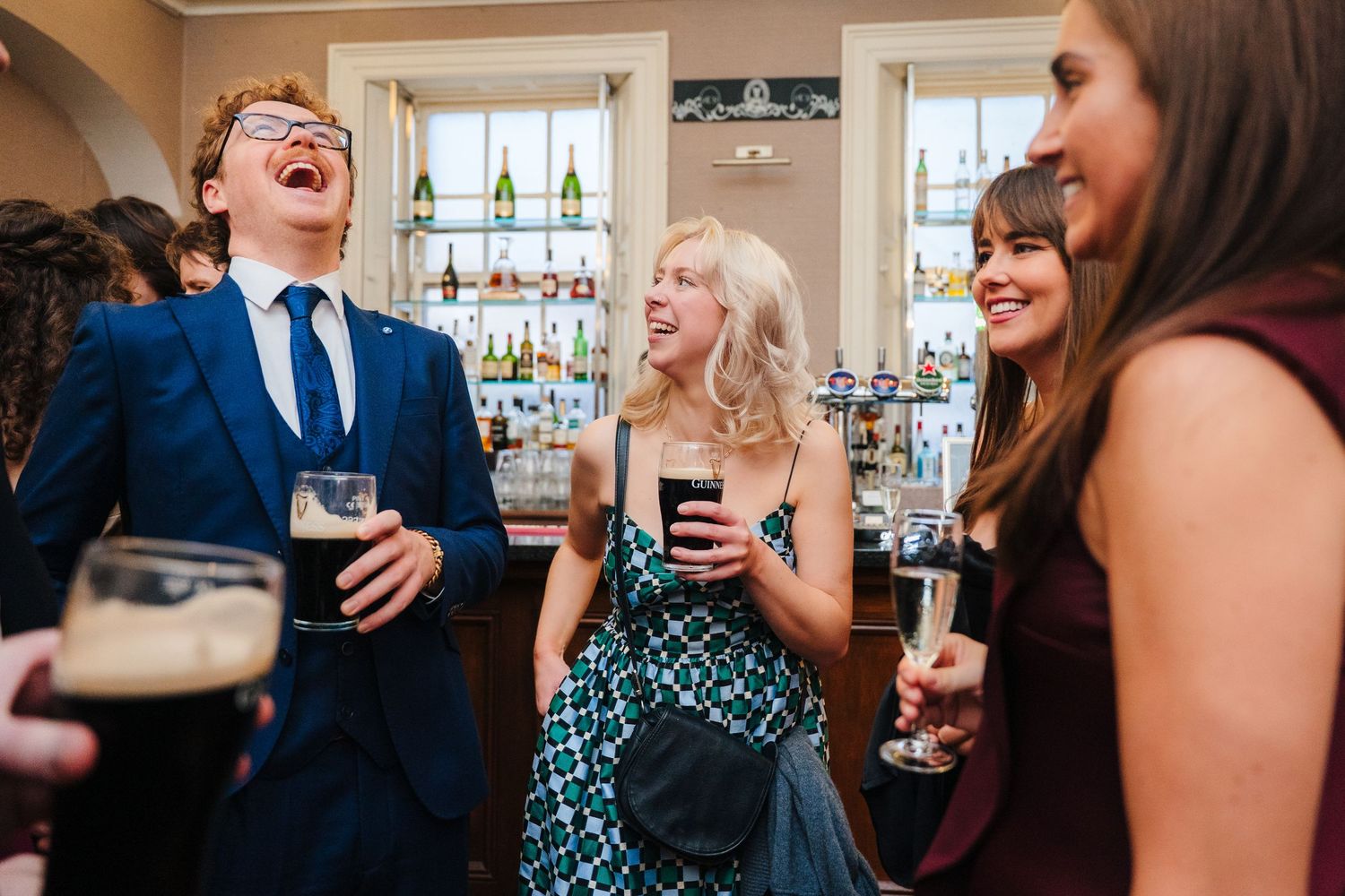 Wedding reception guests laugh and socialize at an elegant indoor venue with large windows. Stephens green club dublin Wedding photographer