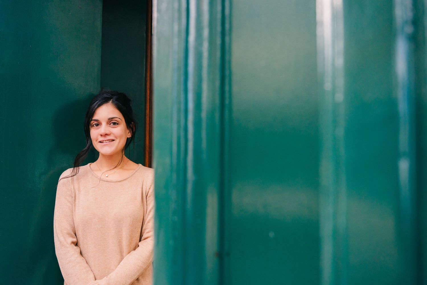 A person in a light colored sweater stands against a teal wall while smiling warmly at the camera.