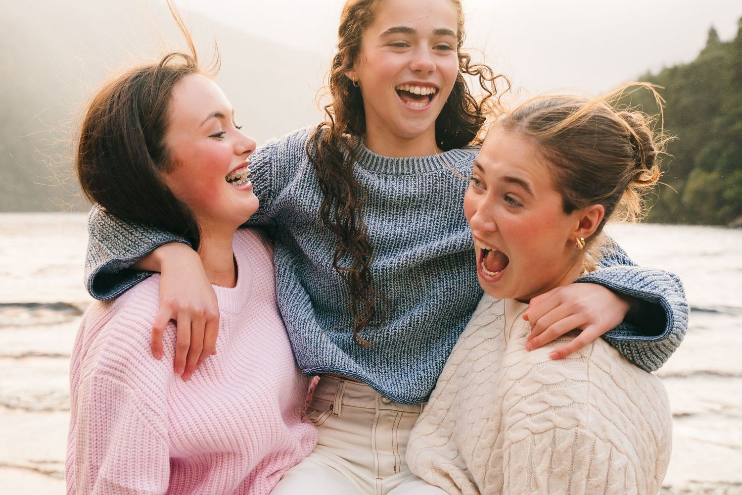 Three friends in cozy sweaters laugh together while enjoying a moment at the beach during sunset. family photographer dublin