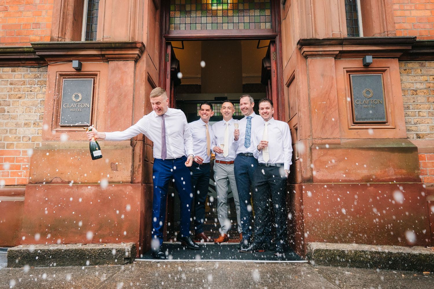 Wedding party spraying champagne outside a brick building in candid celebration shots.