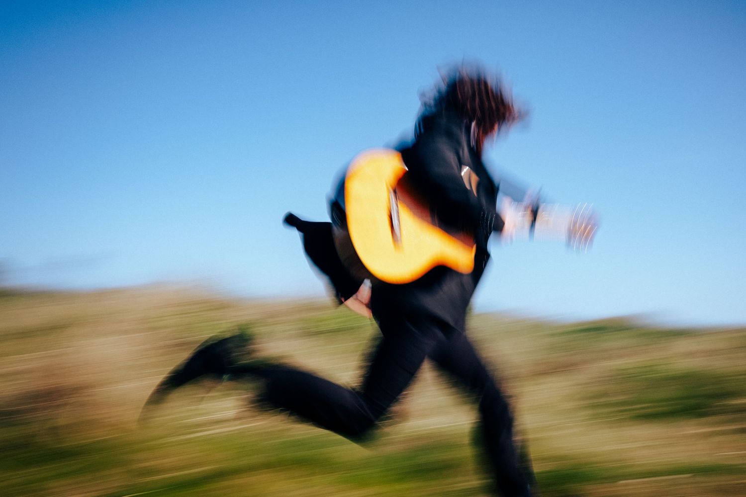 Motion-blurred figure running outdoors in black clothing while carrying an acoustic guitar against blue sky and hills. PHOTOGRAPHER Dublin