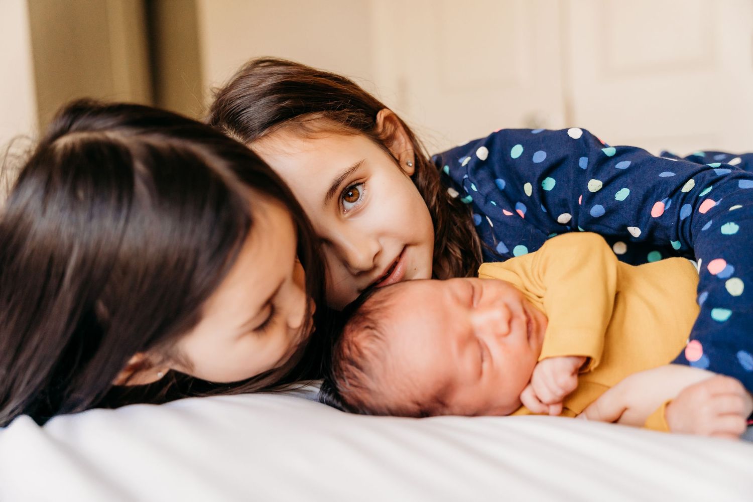 Two older children snuggling close to sleeping baby in yellow onesie on white bed. family PHOTOGRAPHER Dublin