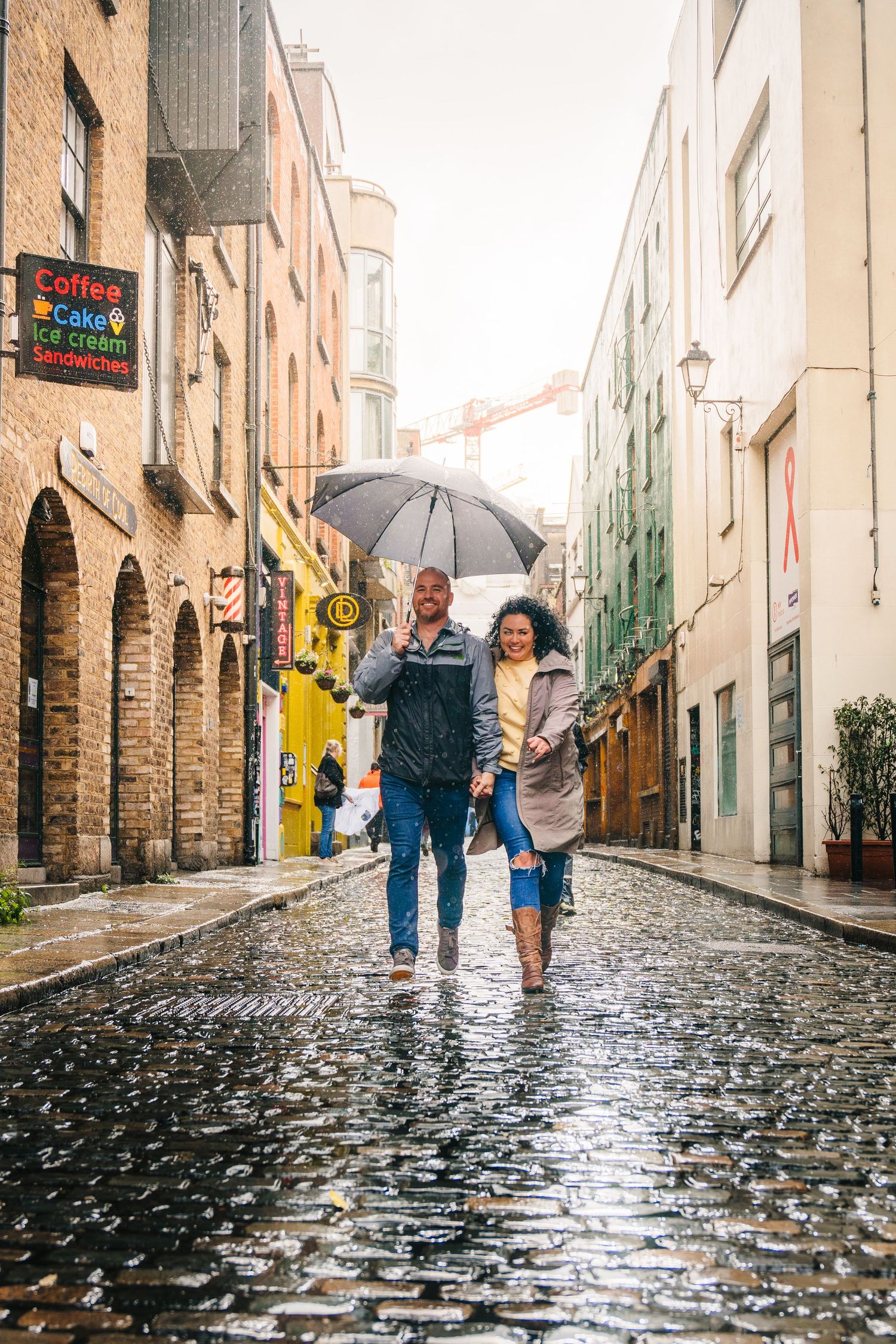 Couple walking together with umbrellas on a wet cobblestone street in a European city during rainfall. Proposal Dublin Photographer