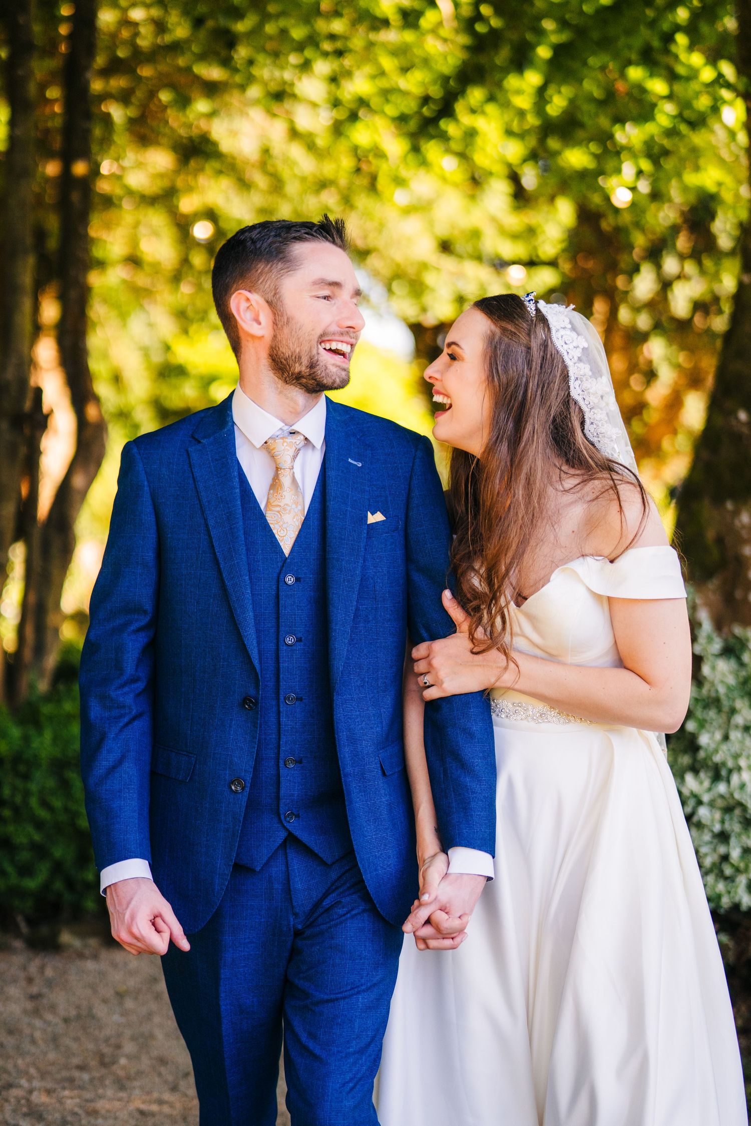 Poulaphouca Wedding photographer wicklow  Newlyweds share joyful moments in a sun-dappled forest setting wearing blue suit and white dress.