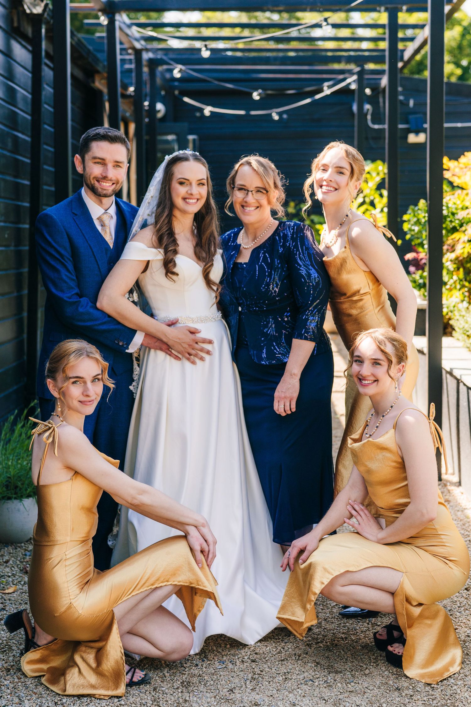 Poulaphouca Wedding pWedding party poses together under a pergola with string lights while wearing gold bridesmaid dresses and a white gown.