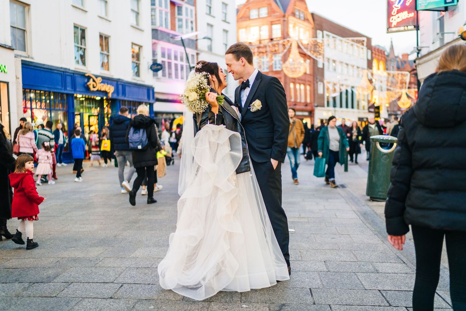 A couple in wedding attire share a moment on a busy city street with shops and pedestrians. Wedding PHOTOGRAPHER Dublin