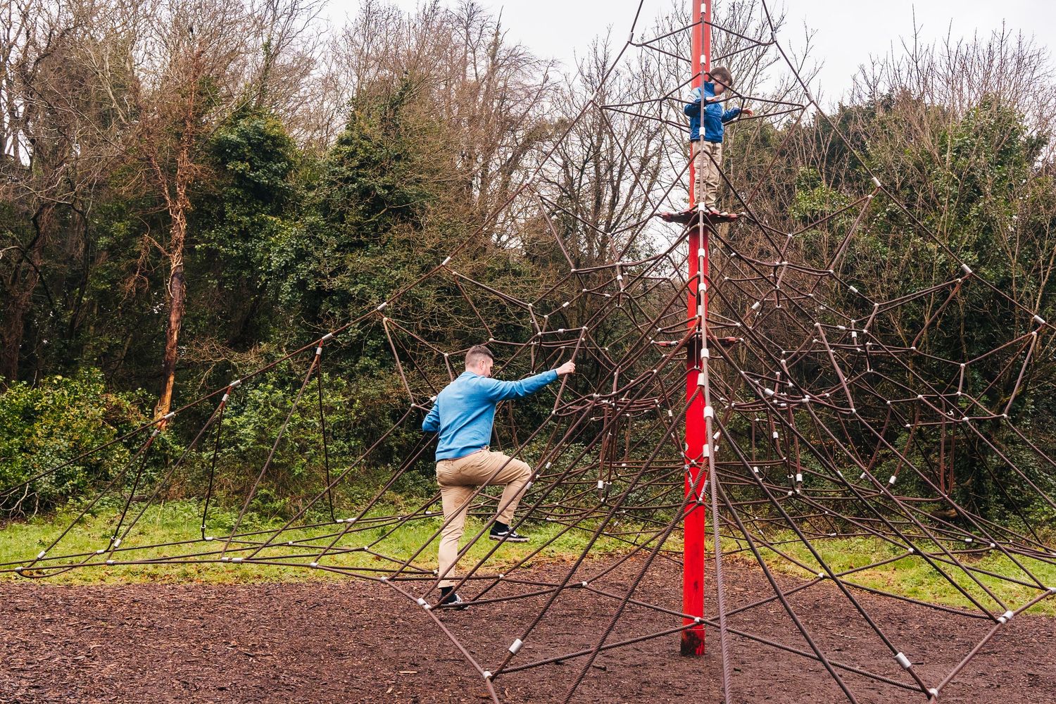 Family photographer Dublin Ireland  Someone in blue climbing on red playground equipment in a wooded park setting.