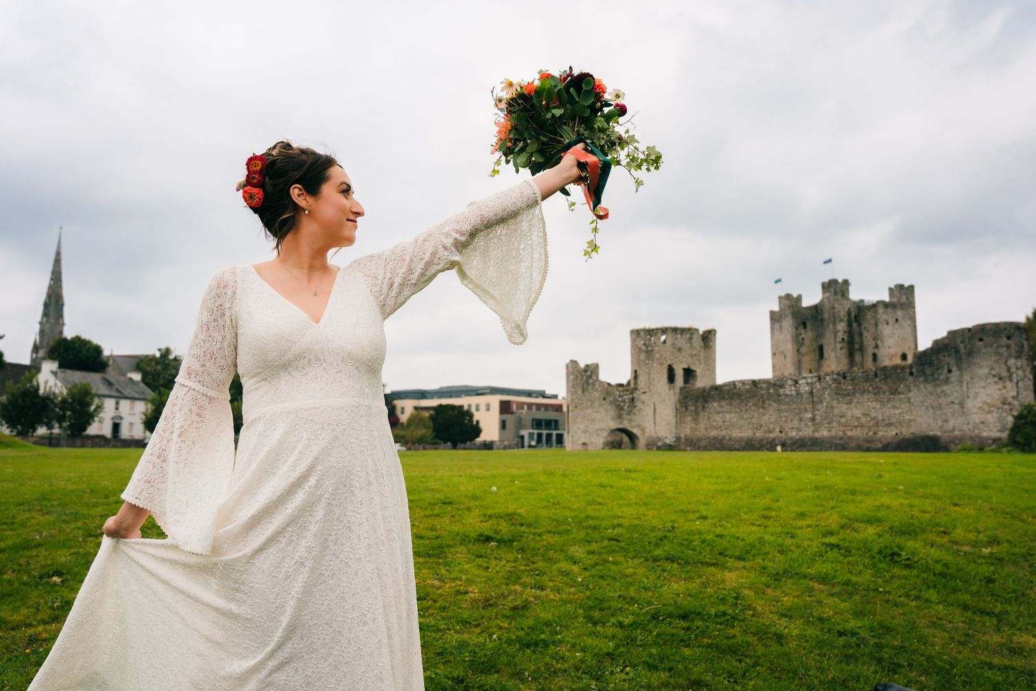 Bride in flowing lace dress holds bouquet aloft against castle backdrop on green lawn.Wedding Photography Trim Castle Ireland