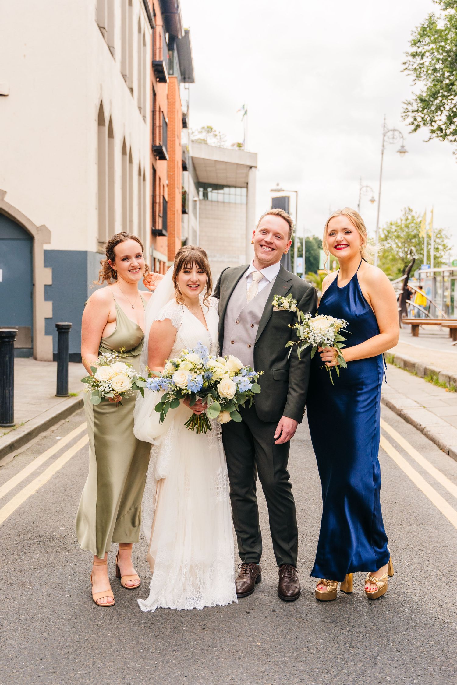 Wedding party poses playfully on a city street wearing formal attire and holding bouquets with white flowers. PHOTOGRAPHER Dublin Ireland We