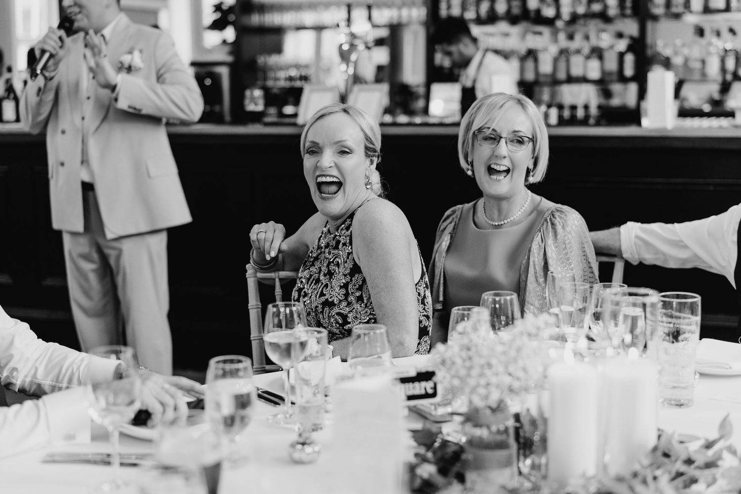 A black and white photo of wedding guests laughing at a reception table. PHOTOGRAPHER Dublin Ireland Wedding